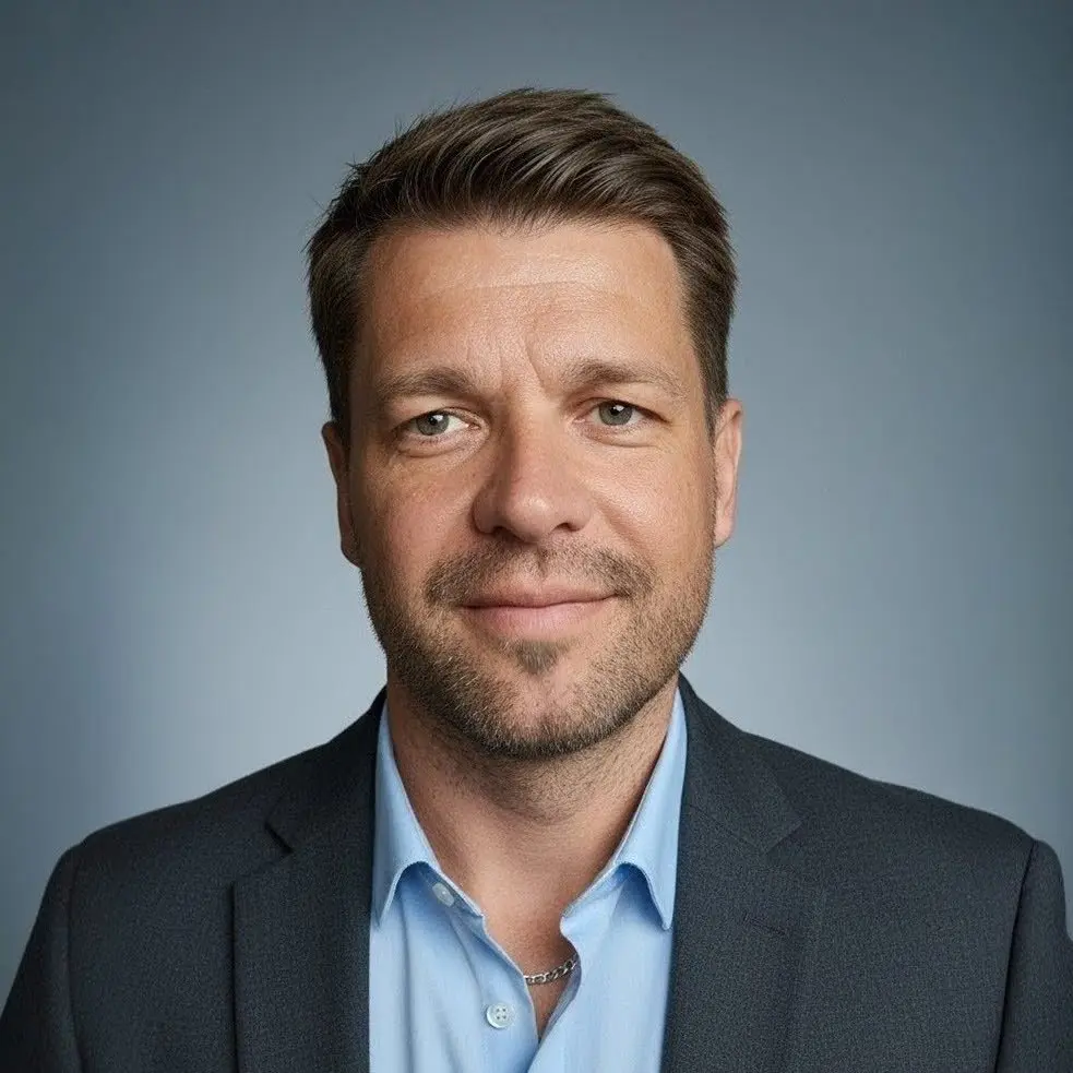 Portraitbild von Roman Neuhäuser als Ausschnitt von Kopf- und Brustbereich. Er lächelt mit geschlossenem Mund, trägt ein hellblaues Hemd (oberer Kopf geöffnet) und ein graues Jacket.