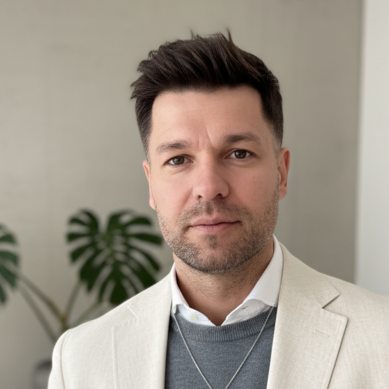 Portraitbild von Roman Neuhäuser als Ausschnitt von Kopf- und Brustbereich. Er lächelt geschmeidig mit geschlossenem Mund, trägt einen grauen Pullover unter dem ein weißer Hemdkragen hervorgeht, mit einem beige-weißem Jackett.