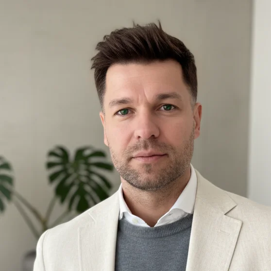 Portraitbild von Roman Neuhäuser als Ausschnitt von Kopf- und Brustbereich. Er lächelt geschmeidig mit geschlossenem Mund, trägt einen grauen Pullover unter dem ein weißer Hemdkragen hervorgeht, mit einem beige-weißem Jackett.